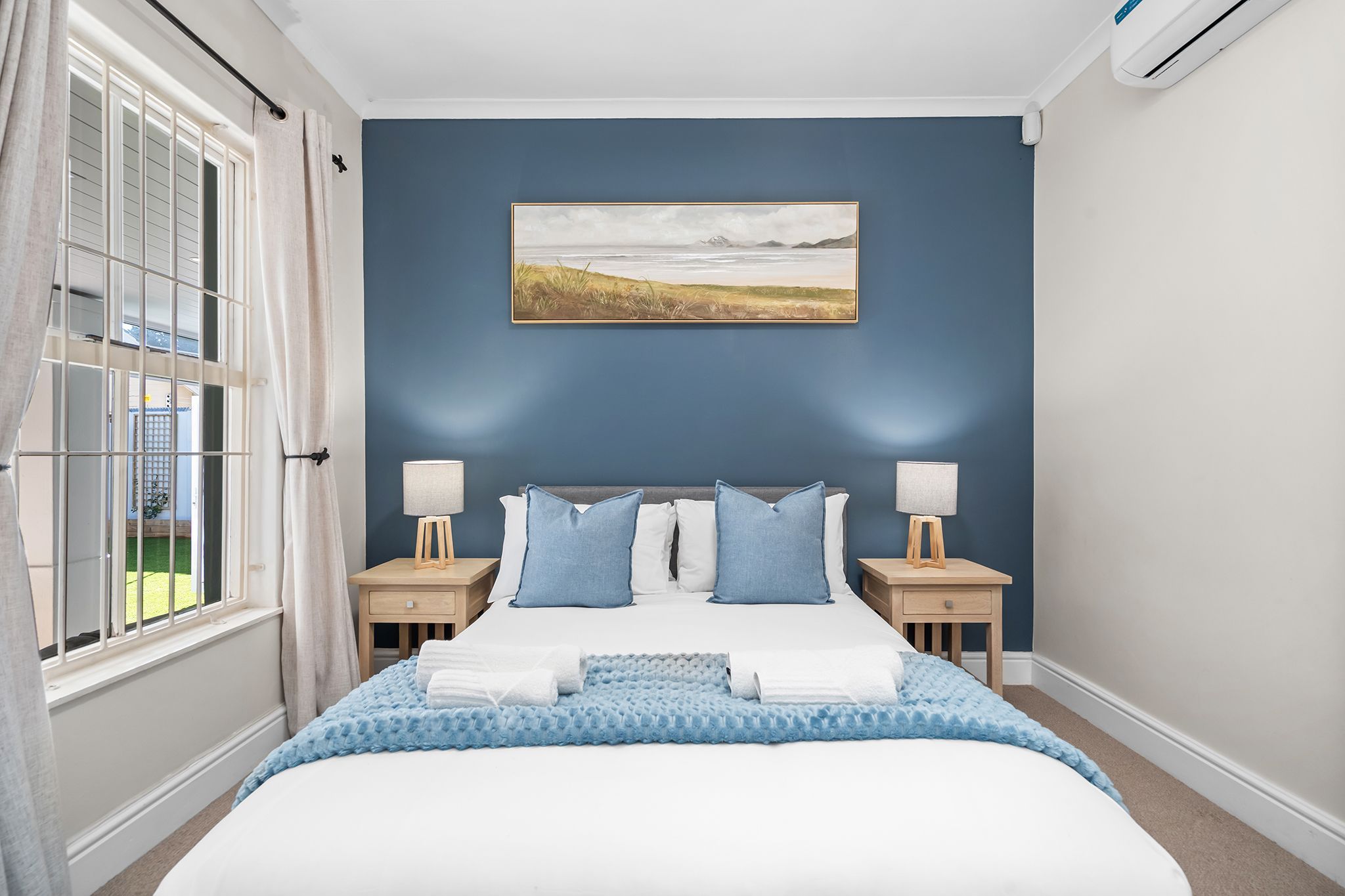 A bedroom with a bed, two bedside tables, a nightstand, a window with curtains, and a wall-mounted painting.