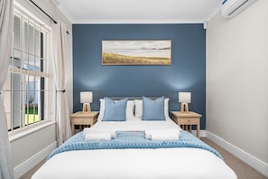 A bedroom with a bed, two bedside tables, a nightstand, a window with curtains, and a wall-mounted painting.