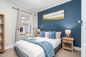 A bedroom with a bed, a window with curtains, a nightstand with a lamp, and a painting on the wall.