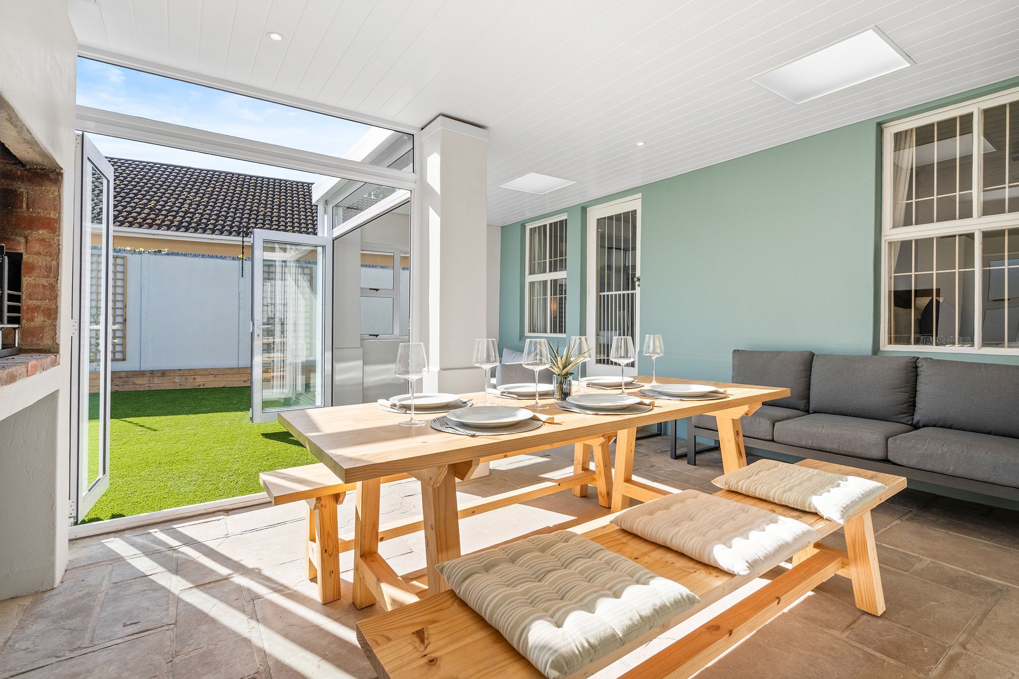 A dining area with a wooden table set for four, a grey sofa, and a view of a green lawn through glass doors.