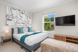 Un dormitorio moderno con una cama grande, una mesita de noche con lámpara, un televisor montado en la pared y una ventana con vistas a zonas verdes.