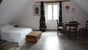 Een slaapkamer met een bed, een houten kist, een raam met gordijnen, een tafel met stoelen en een wand met decoratieve objecten.