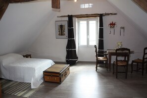 A bedroom with a bed, a wooden chest, a window with curtains, a table with chairs, and a wall with decorative items. - Le Jardin aux Lilas – Private Pool, Renovated House in Lot Valley (Cadrieu)