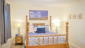 A four-poster bed with a headboard, two bedside tables, a wall-mounted lamp, a painting, and a window with curtains.