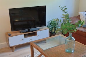 Ein Wohnzimmer mit Flachbildfernseher auf einer Holzeinrichtung, einem Glastisch, einer Topfpflanze und einem braunen Ledersofa.