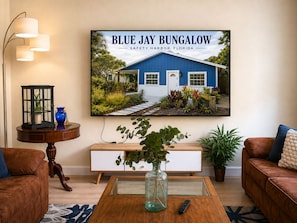 Un salon comprenant un grand tableau encadré représentant une maison de style bungalow, une table basse en bois, un canapé en cuir marron, une console blanche et un lampadaire.