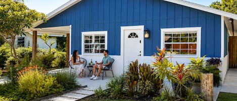 Une maison à deux étages, avec une façade bleue, un porche à l’avant doté de chaises et un jardin où poussent différentes espèces de plantes.