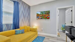 A living room with a yellow sofa, a blue rug, and a painting of a garden on the wall. - 2 Bed Oasis 10 Minutes to Downtown (tampa)