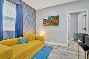 A living room with a yellow sofa, a blue rug, and a painting of a garden on the wall. - 2 Bed Oasis 10 Minutes to Downtown (tampa)