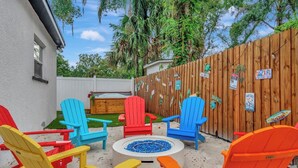 Un patio trasero con cerca de madera, sillas Adirondack de colores, un hoyo para fuego y una terraza de madera.