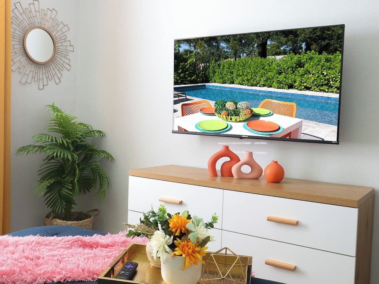 Un salon moderne avec un téléviseur fixé au mur, un meuble en bois agrémenté d’objets décoratifs, un tapis rose et moelleux, et une plante en pot.