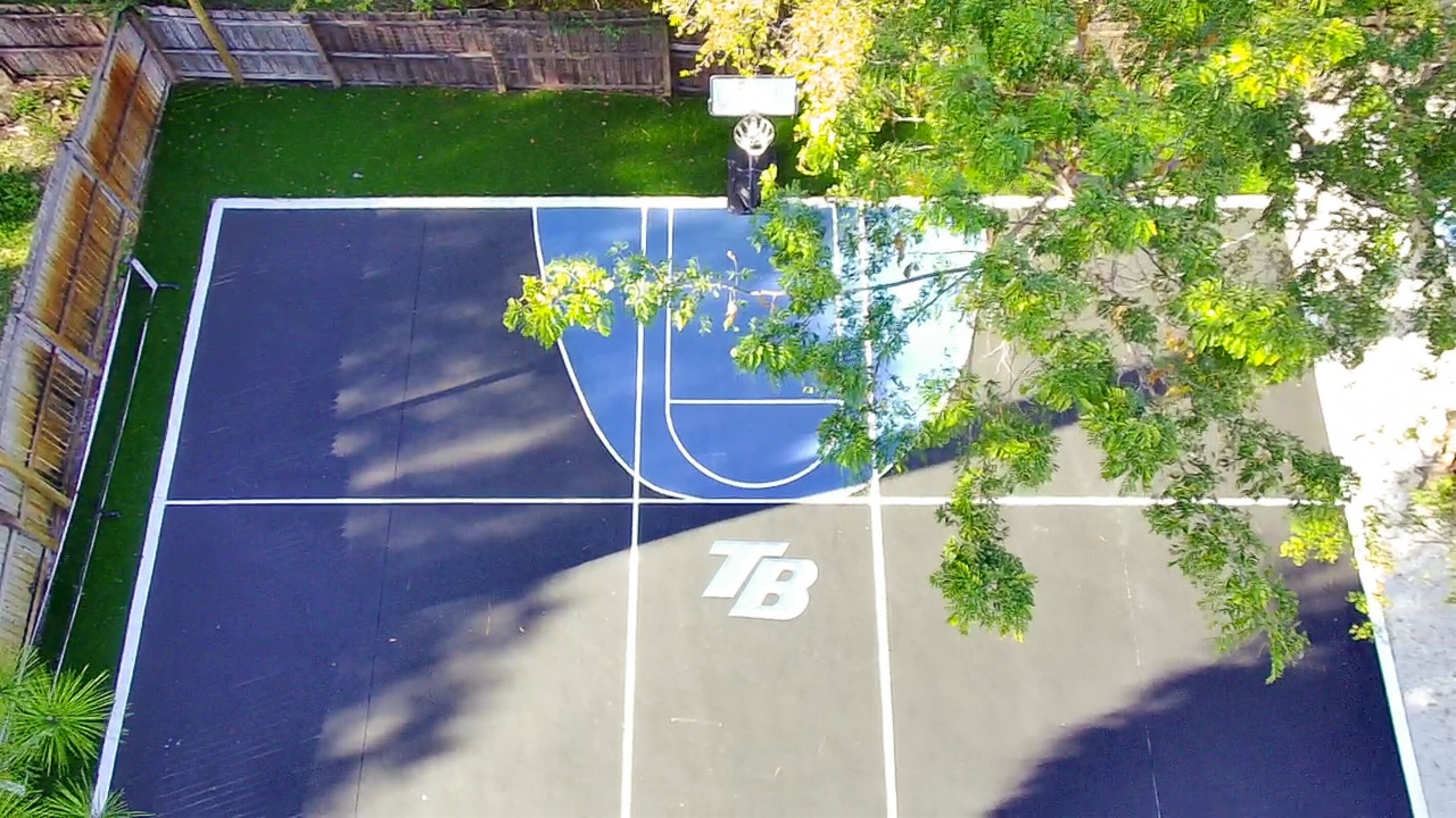 An outdoor basketball court with a 'TB' marking, surrounded by greenery and a wooden fence.