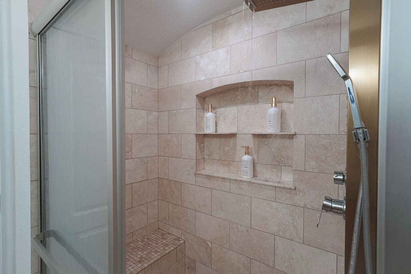 A shower with a glass door, beige tiles, and a shelf with toiletries.