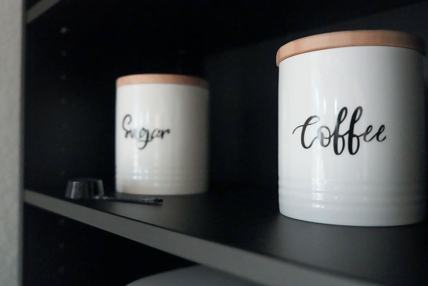 To hvide beholdere med låg står på en hylde. Den ene beholder er mærket 'Sugar', og den anden 'Coffee'.