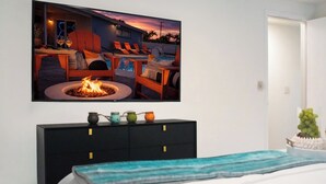 A bedroom with a bed, a black dresser with colorful jars, a painting of a beach scene, and a television displaying a poolside scene. - Tampa High Roller, Relax and Play (Tampa)