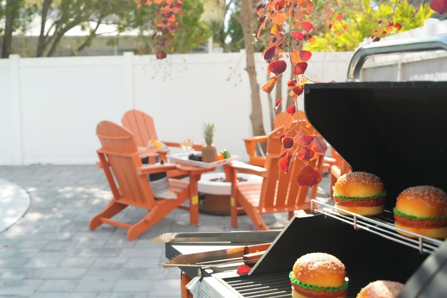 An outdoor dining area with wooden chairs and a table, a grill with burgers, and hanging plants.