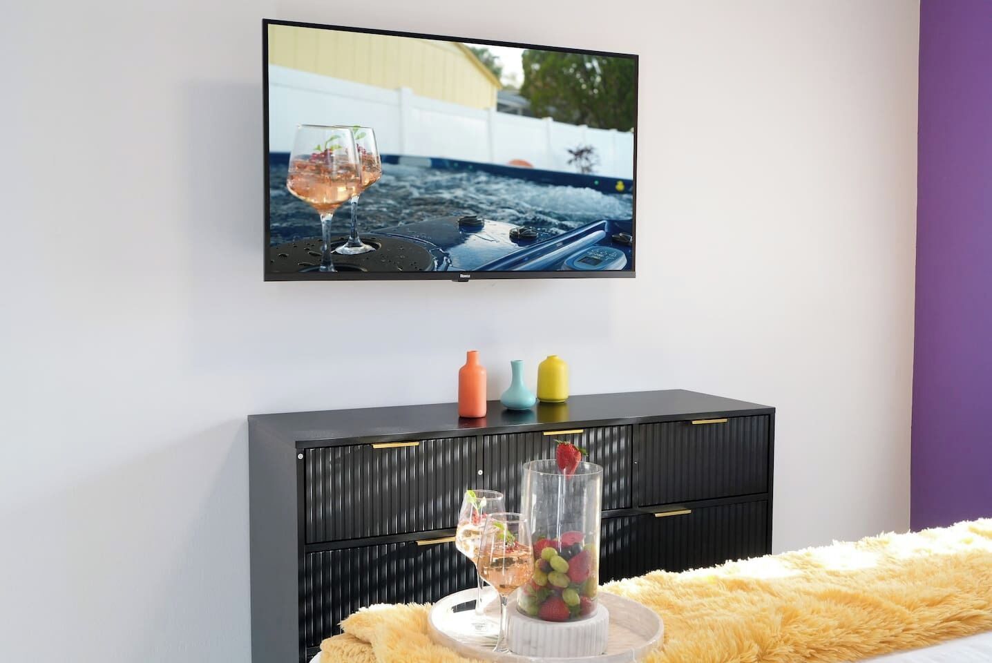 A modern living room with a flat-screen TV mounted on the wall, a black cabinet with colorful vases, and a fluffy yellow throw on the sofa.
