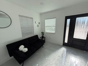 A modern living room with a black sofa, a round mirror, and two windows with blinds. - Mini Home in Tampa for Two (Tampa)