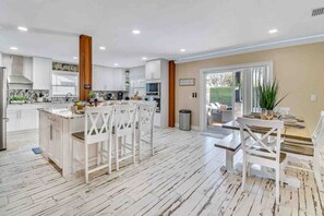 A modern kitchen with white cabinetry, a central island with chairs, and a dining area with a table and chairs. - Villa 5 Bed With Saltwater Heated Pool & Jacuzzi (tampa)