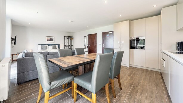 A modern kitchen with a built-in oven and a dining area with a wooden table and grey upholstered chairs. - Cotswolds Apartment 2 Sleeps 4 (Chipping Norton)