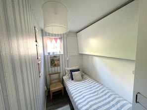 Ein kleiner, schmaler Raum mit einem Einzelbett, einem Stuhl, einem Fenster mit Vorhängen und einem an der Wand befestigten Regal.