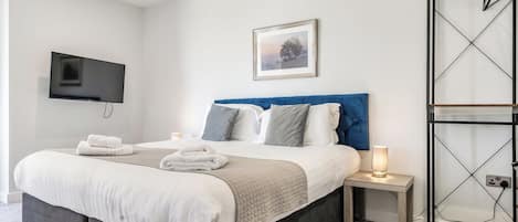 A modern hotel room with a bed, bedside tables, a TV, a framed picture, and a wardrobe.