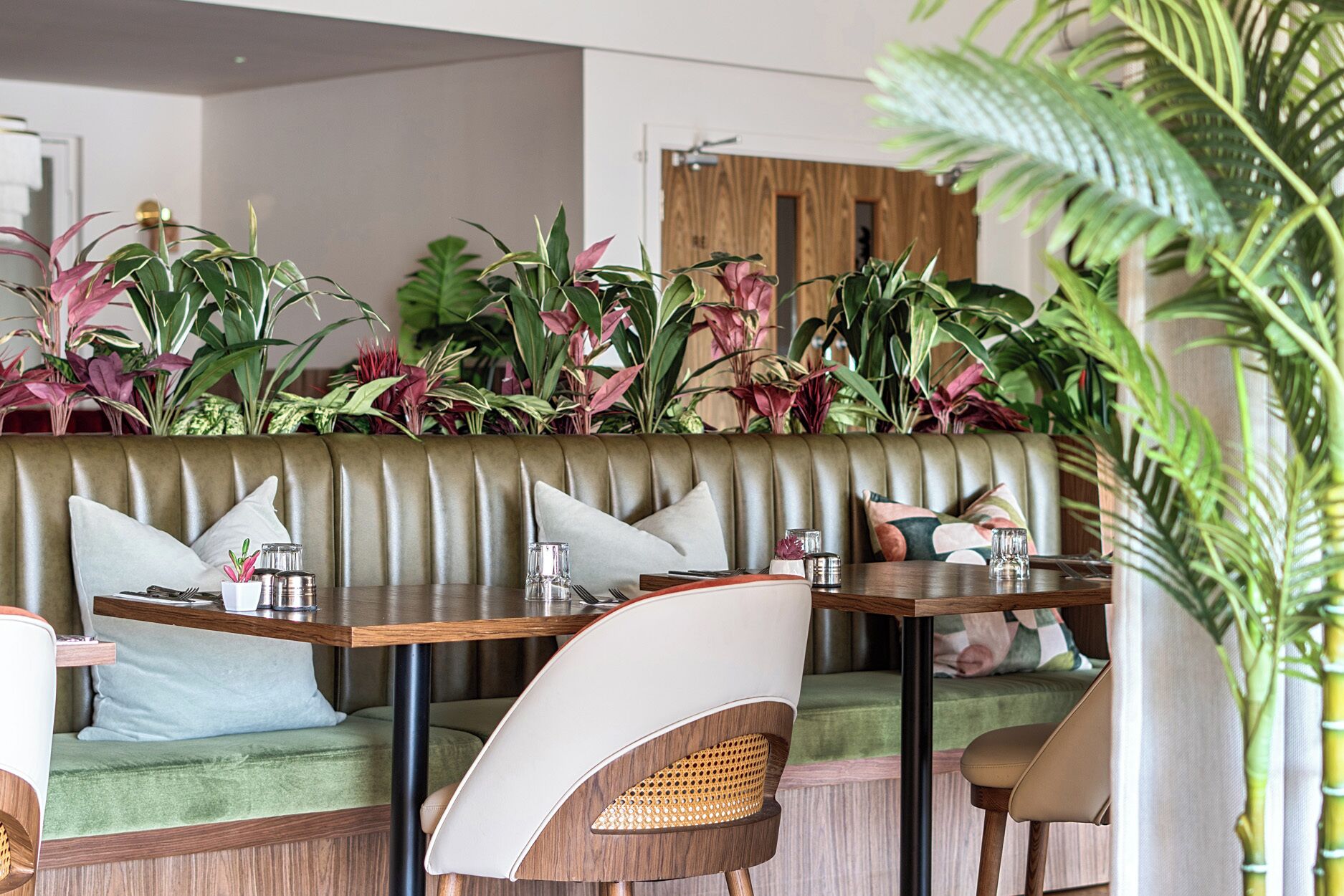 A restaurant with a booth seating area, wooden tables, and a variety of plants.