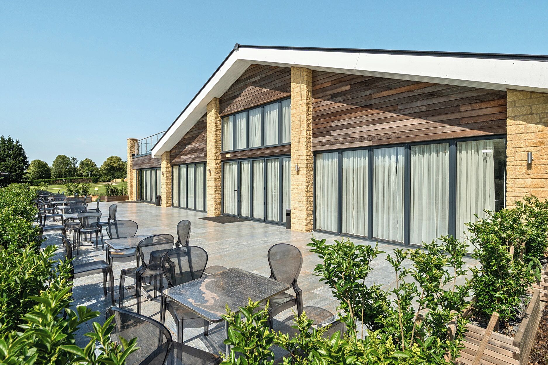 Un espace extérieur moderne, aménagé avec des tables et des chaises, doté d’une façade en pierre et de grandes fenêtres.