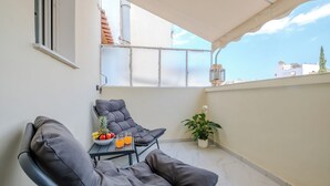 Un balcon avec deux fauteuils de relaxation noirs, une petite table avec des fruits et des boissons, une plante en pot et une vue sur des immeubles et un ciel dégagé.