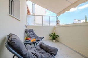 A balcony with two black lounge chairs, a small table with fruits and drinks, a potted plant, and a view of buildings and a clear sky. - Academy Stay Athens | Modern Apartment Near Metro (Athina)