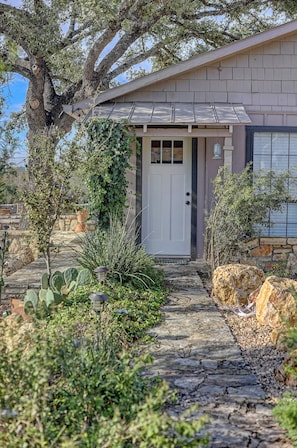 A house with a stone pathway leading to a white door, surrounded by various plants and a large tree. - Hunt River Retreat Porch, Kayaks & Hill Country (Hunt)