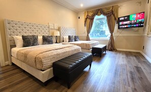Ein Hotelzimmer mit einem großen Bett, einem kleineren Bett, einer Bank, einem Fernseher und einem Fenster mit Vorhängen.