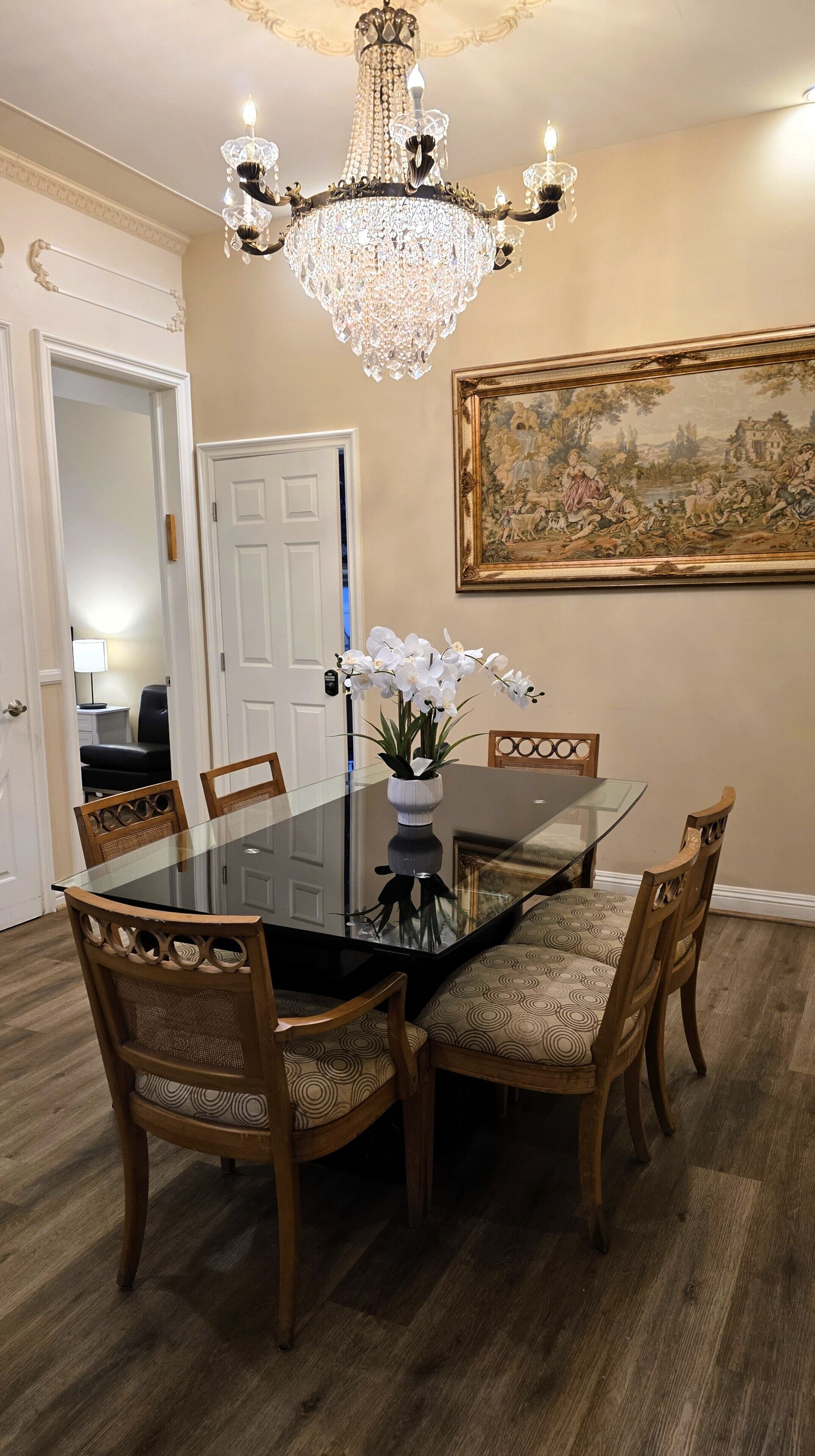 A dining room with a glass table, wooden chairs, a chandelier, and a landscape painting on the wall.