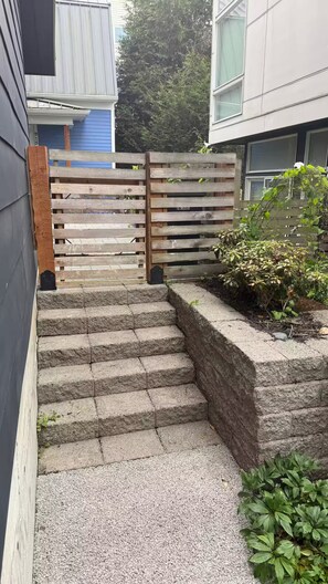 A set of stone steps leading to a wooden gate, surrounded by greenery and a building with a blue exterior. - The May Townhome - Lower (Seattle)