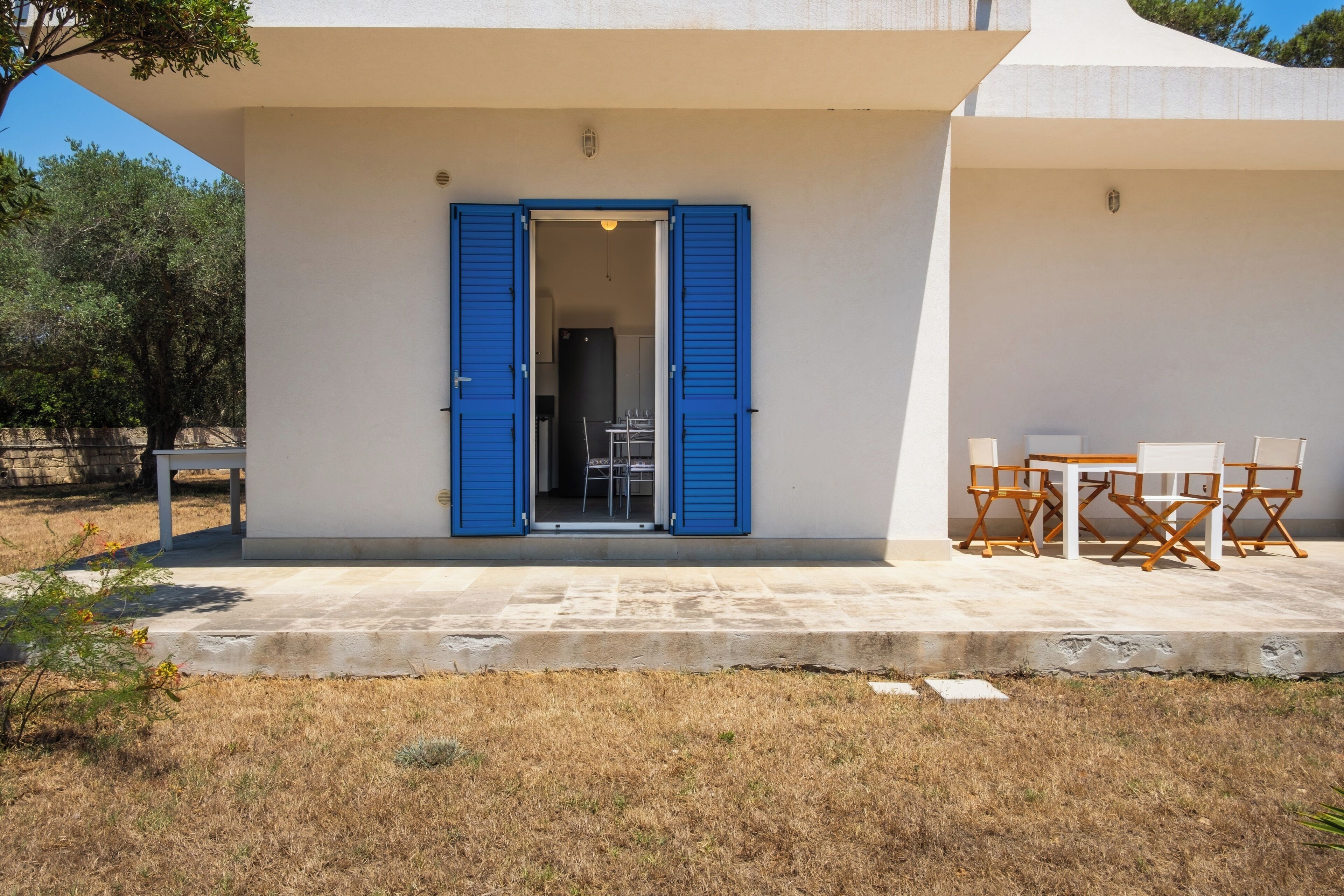 Ein modernes Gebäude mit weißer Fassade und blauen Fensterläden. Eine Terrasse mit Gartenmöbeln ist vorhanden.