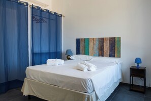 Een tweepersoonsbed met witte lakens, een nachtkastje met een lamp en een blauw gordijn.