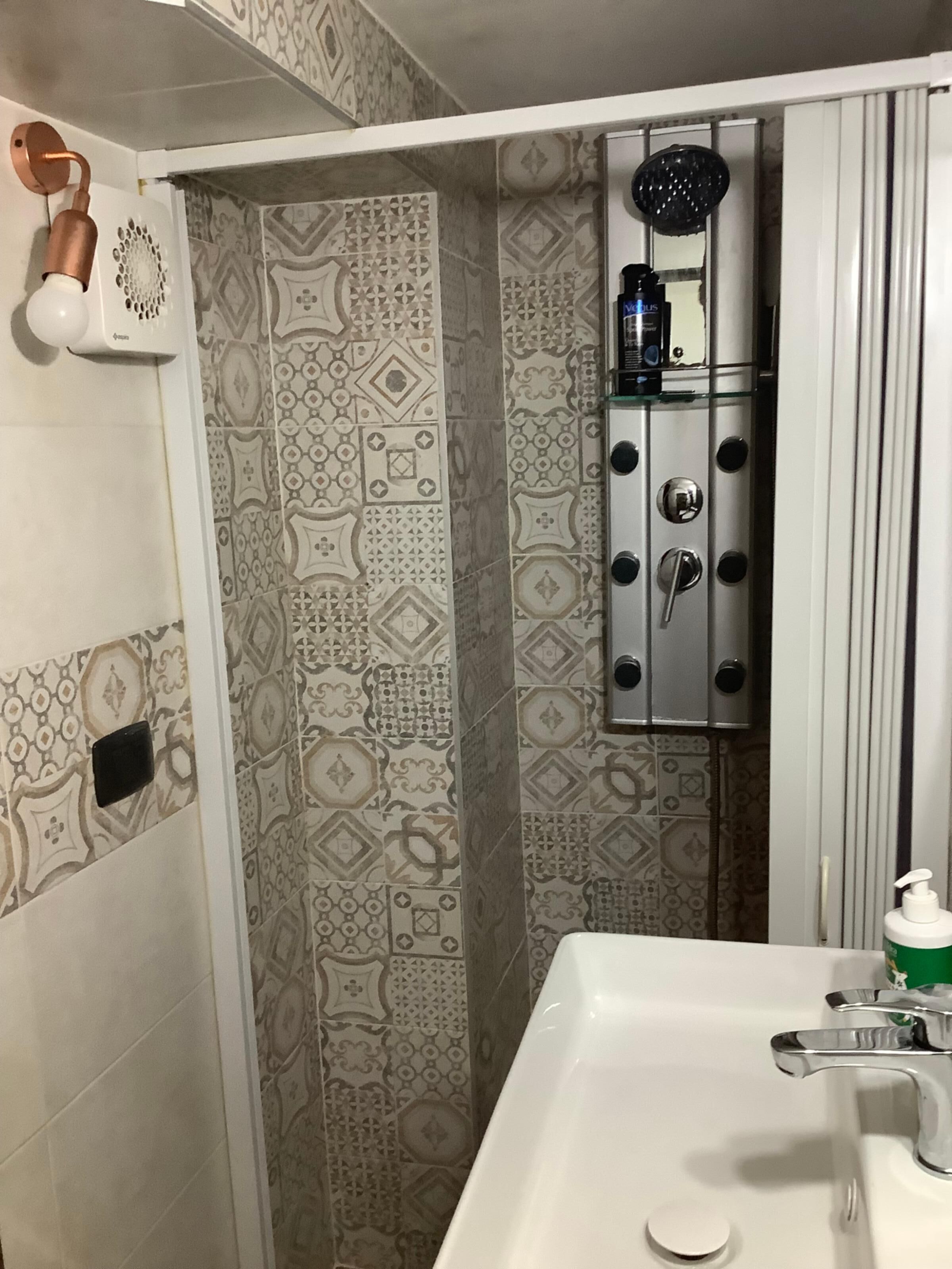 Un baño con baldosas estampadas, un lavamanos blanco, un cabezal de ducha de mano y una ducha con mampara de vidrio y dispensador de jabón.