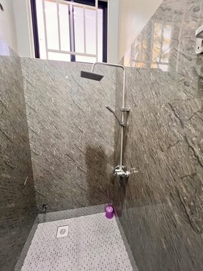 A shower area with a rain showerhead and handheld showerhead, a drain, and a purple object on the floor.