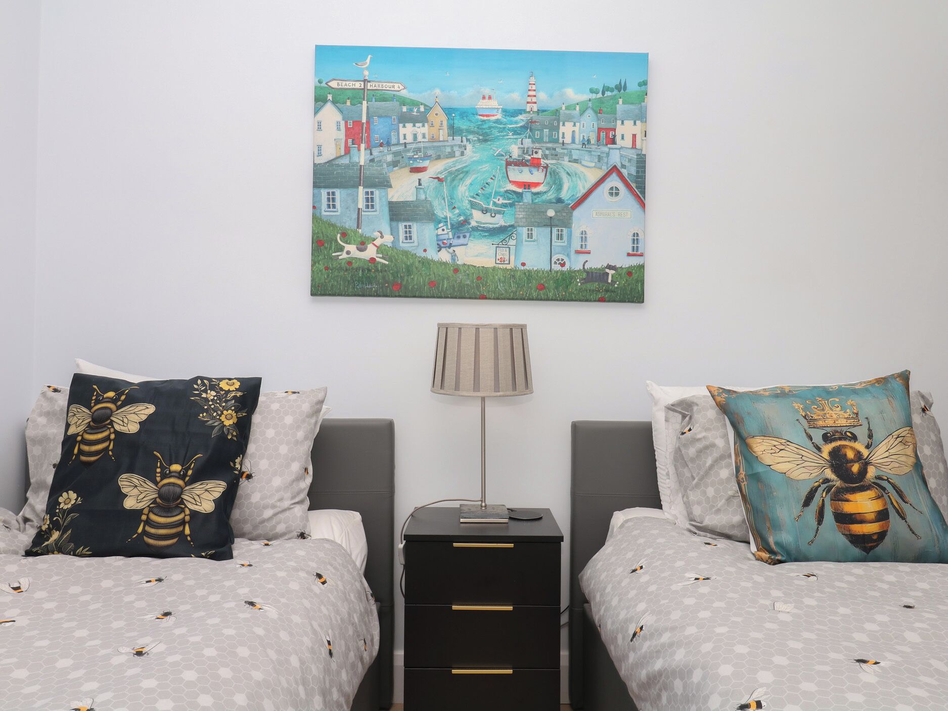 A bedroom with two beds, a nightstand, and a painting of a coastal town.
