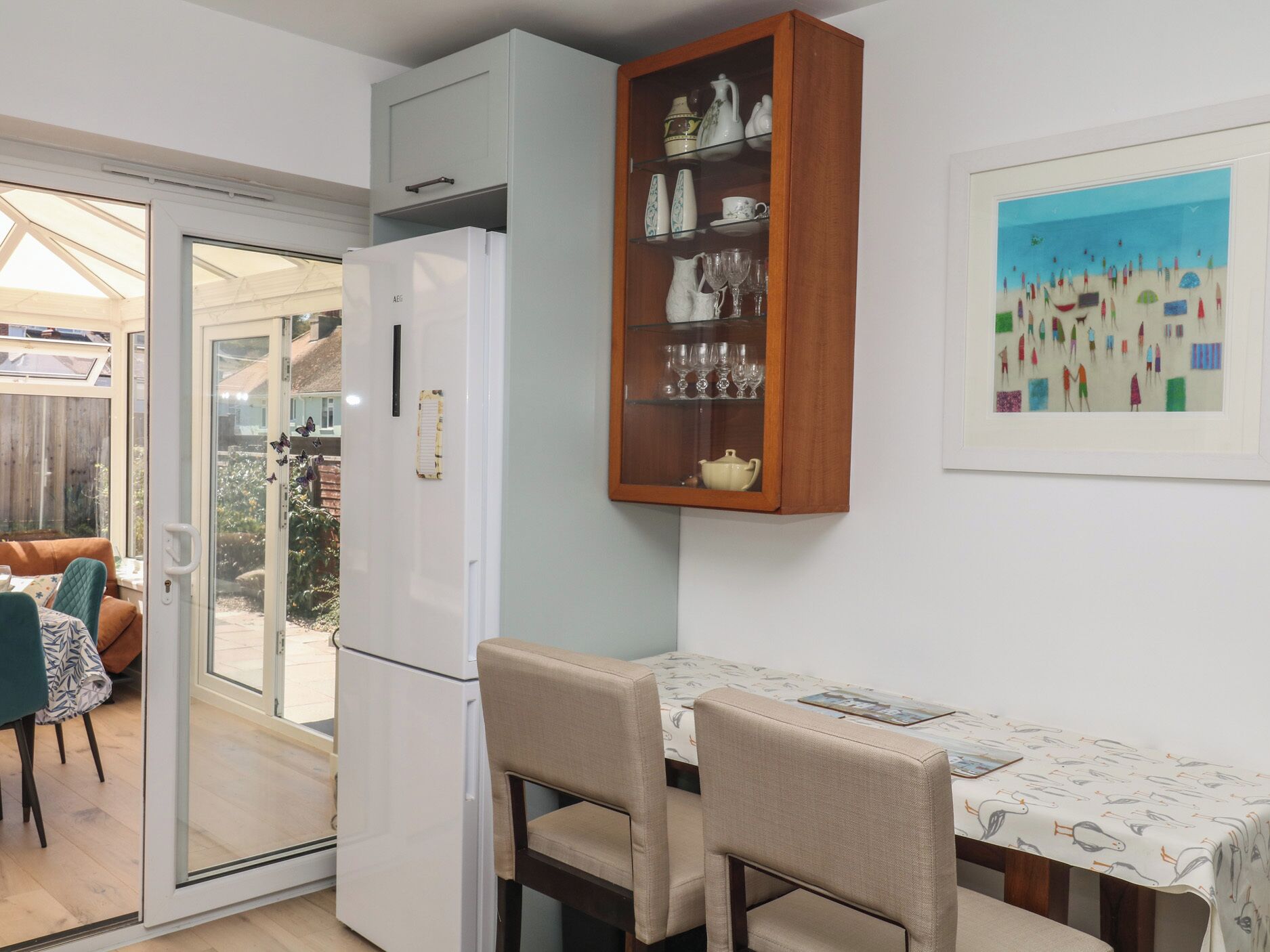 A compact kitchen with a refrigerator, a wooden cabinet with glass shelves, a dining table with chairs, and a framed artwork on the wall.