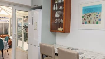 A compact kitchen with a refrigerator, a wooden cabinet with glass shelves, a dining table with chairs, and a framed artwork on the wall.