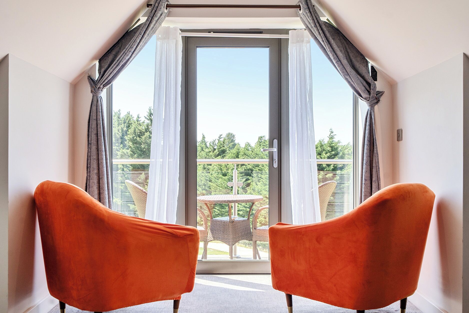 A room with two orange chairs facing a balcony with a view of greenery.
