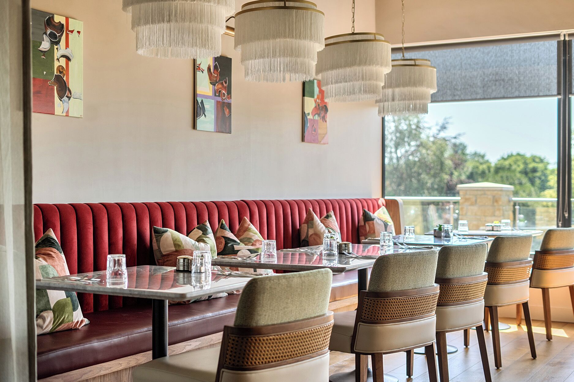 A restaurant with a red upholstered booth, multiple tables with chairs, hanging chandeliers, and artwork on the walls.