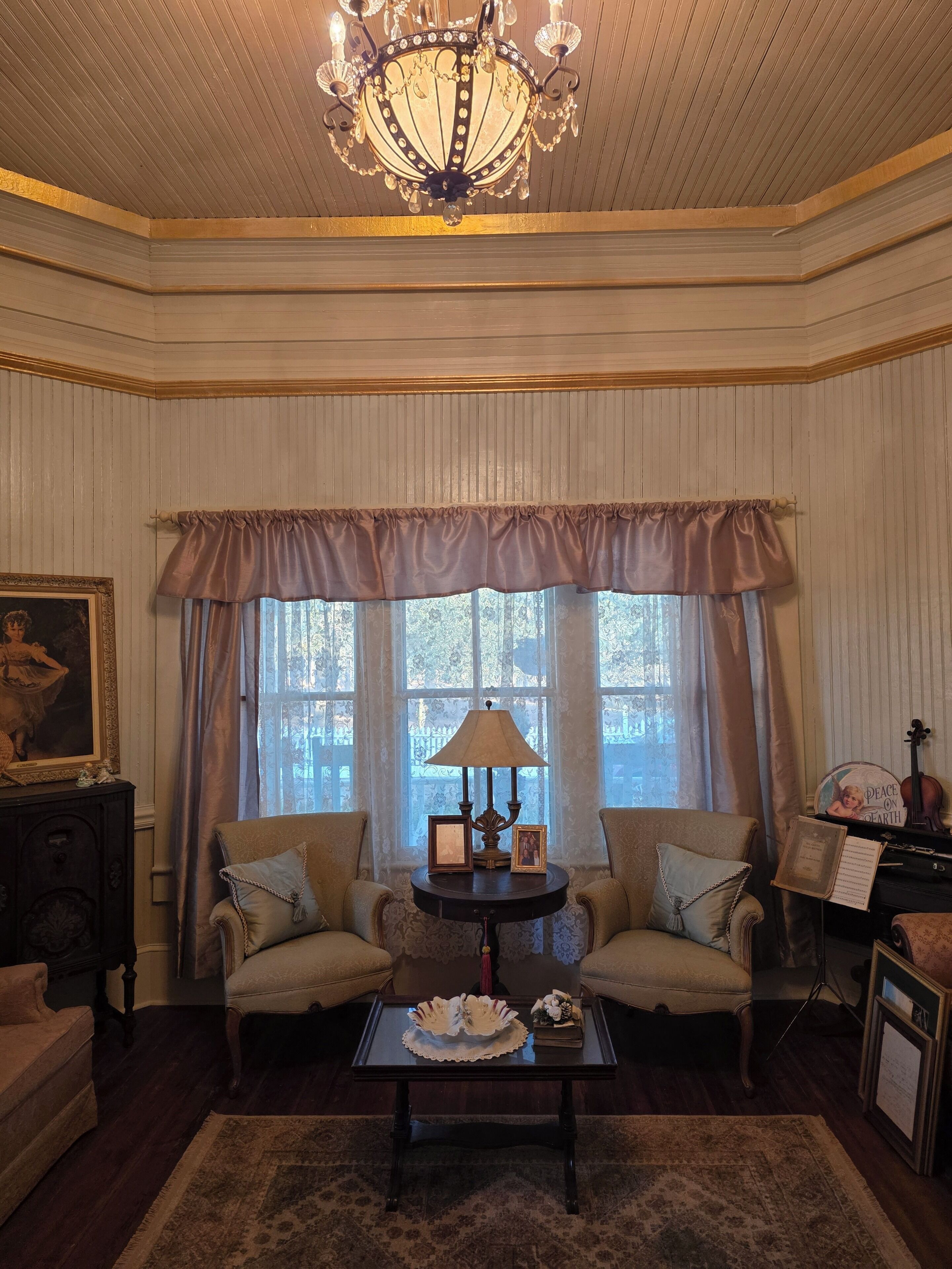 A room with a chandelier, two armchairs, a table with a lamp, and a piano.