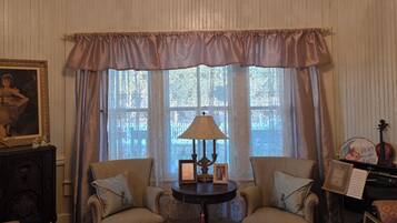 A room with a chandelier, two armchairs, a table with a lamp, and a piano.