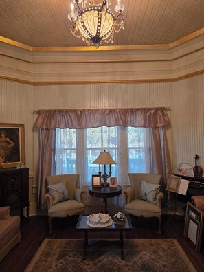 A room with a chandelier, two armchairs, a table with a lamp, and a piano.