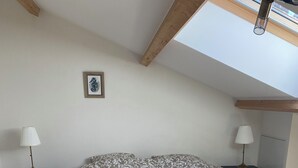 Une chambre avec un grand lit, deux tables de chevet avec des lampes, un tableau encadré au mur et une lucarne au plafond.