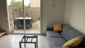 A living room with a grey sofa, two yellow pillows, a glass coffee table, and a sliding glass door leading to an outdoor patio with a table and chairs. - Joalland's Cozy Retreat, 50 meters from the beach (La Plaine-sur-Mer)