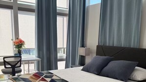 A hotel room with a bed, a desk with a chair, a vase of flowers, and large windows with curtains. - The May Townhome - Upper (Seattle)