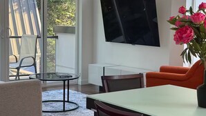 A modern living room with a dining table, chairs, a TV mounted on the wall, and a large window with a view of the outdoors. - The May Townhome - Upper (Seattle)